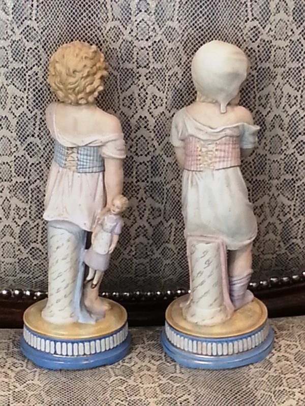 Pareja Figuras de porcelana Biscuit: Niña con muñeca en la mano y Niña con sombrero. S. XIX.